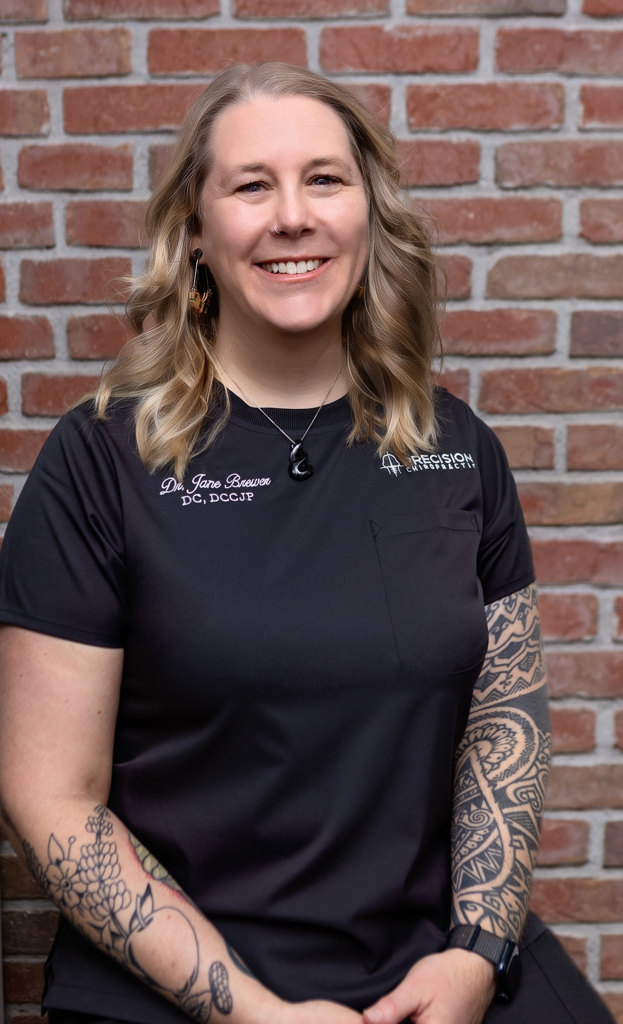 Dr. Jane Brewer, DC DCCJP, seated in Precision Chiropractic scrubs against a brick wall, Strategic Partner of the Combat Recovery Foundation